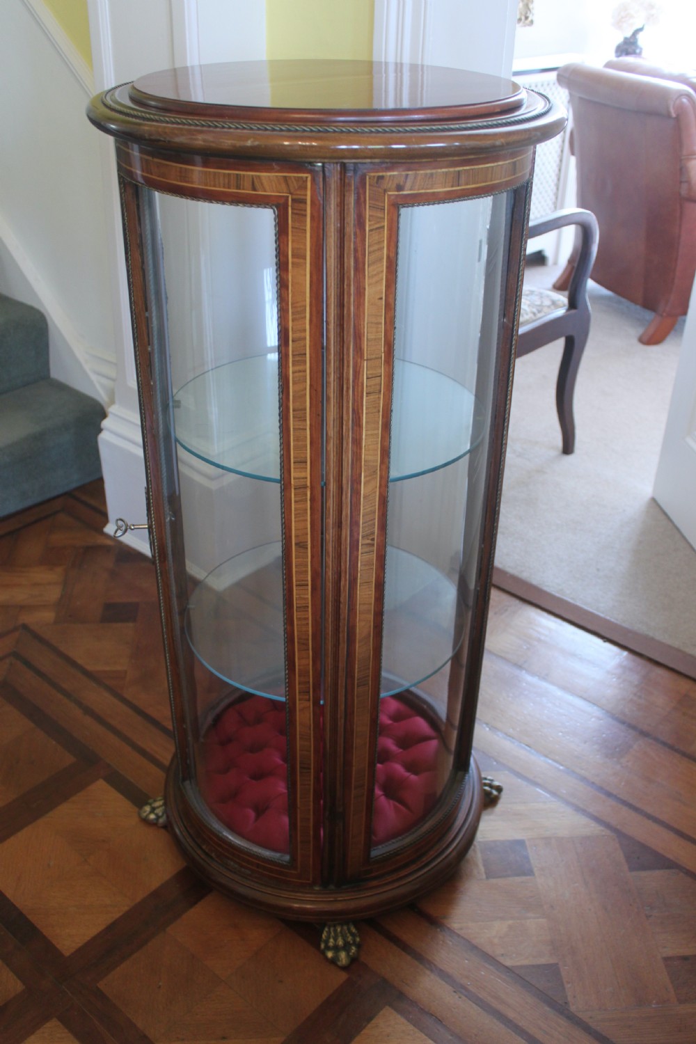 A late 19th-century cylindrical French vitrine cabinet featuring inlaid timber patterning, curved glass panels, tufted red velvet interior lining, and brass claw feet. View V