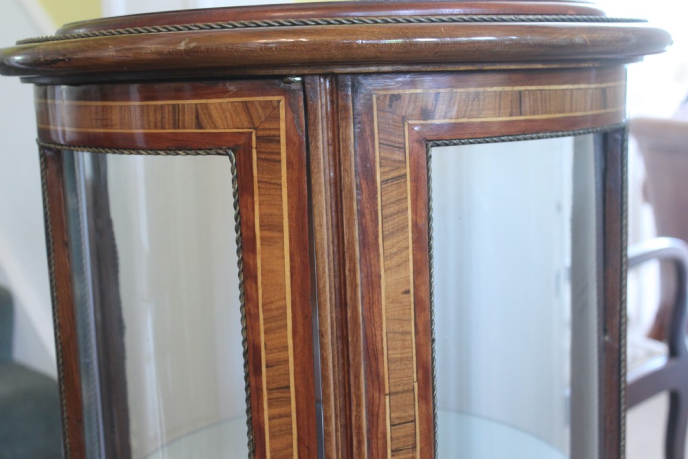A late 19th-century cylindrical French vitrine cabinet featuring inlaid timber patterning, curved glass panels, tufted red velvet interior lining, and brass claw feet. View VII