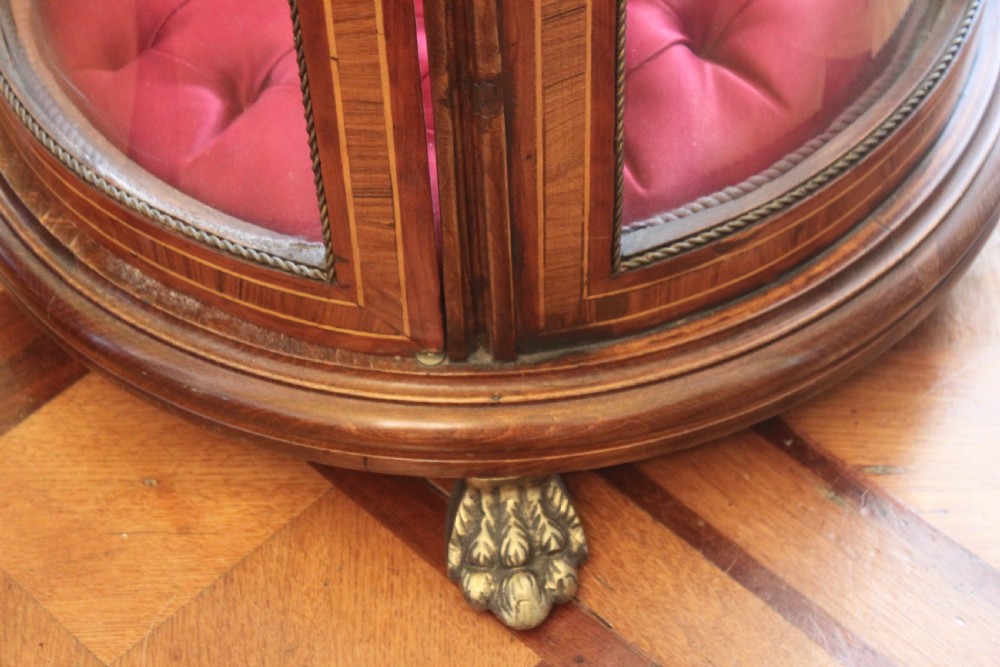 A late 19th-century cylindrical French vitrine cabinet featuring inlaid timber patterning, curved glass panels, tufted red velvet interior lining, and brass claw feet. View VIII