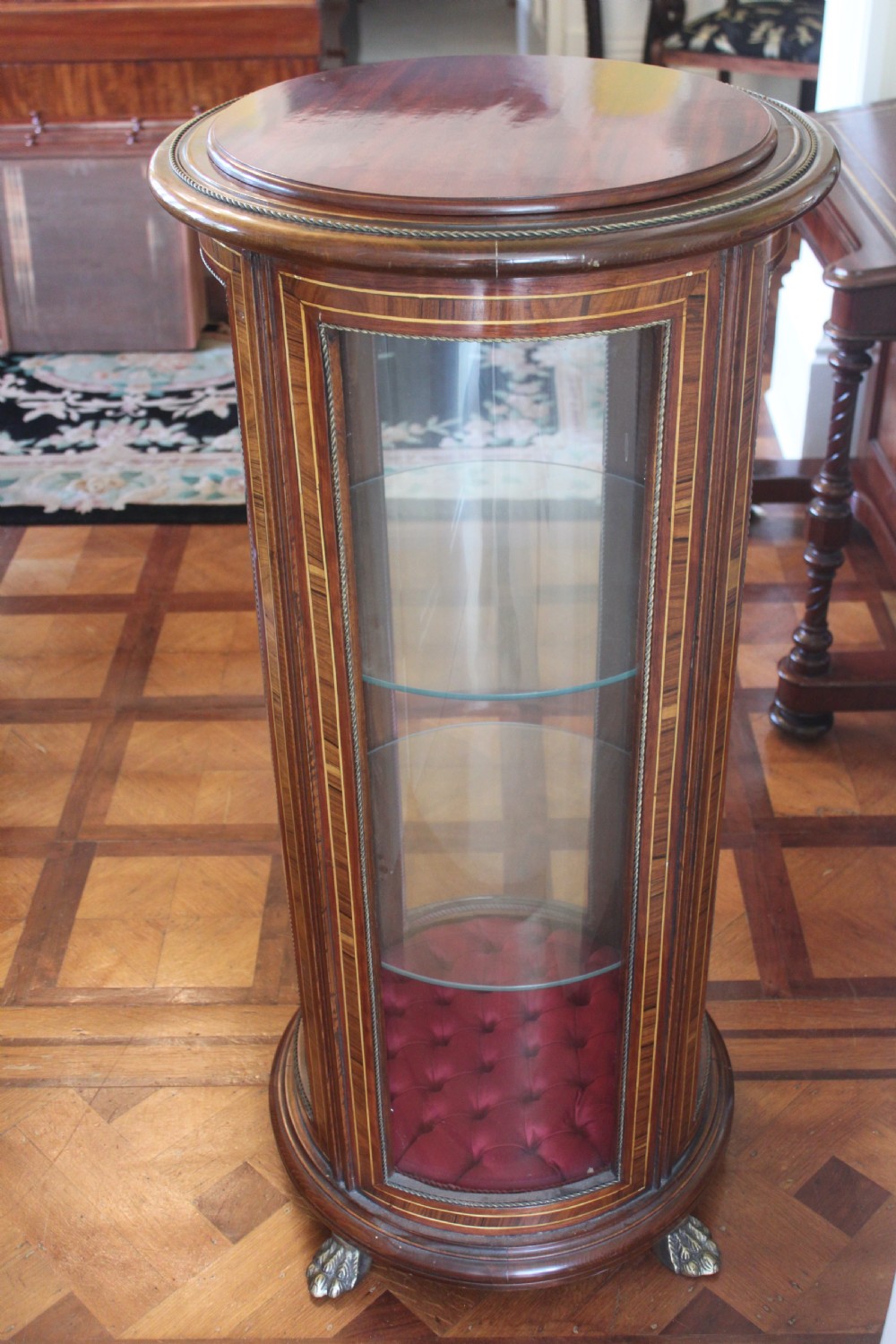 A late 19th-century cylindrical French vitrine cabinet featuring inlaid timber patterning, curved glass panels, tufted red velvet interior lining, and brass claw feet. View IX