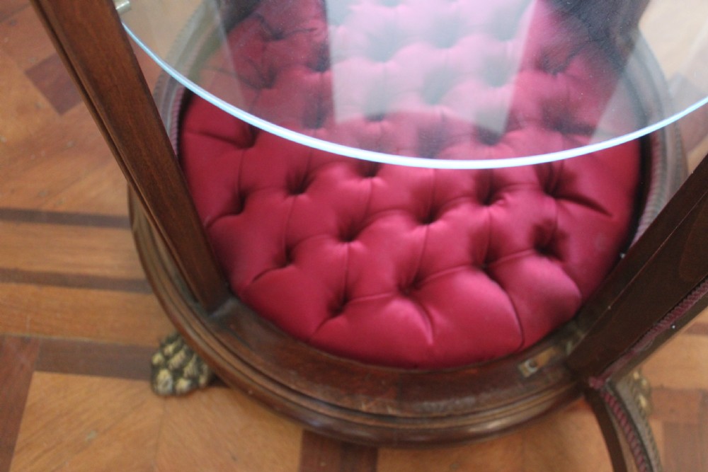 A late 19th-century cylindrical French vitrine cabinet featuring inlaid timber patterning, curved glass panels, tufted red velvet interior lining, and brass claw feet. View X