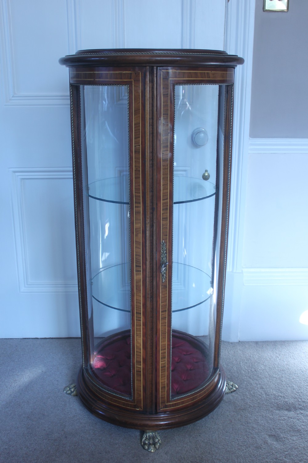 A late 19th-century cylindrical French vitrine cabinet featuring inlaid timber patterning, curved glass panels, tufted red velvet interior lining, and brass claw feet. View XIII