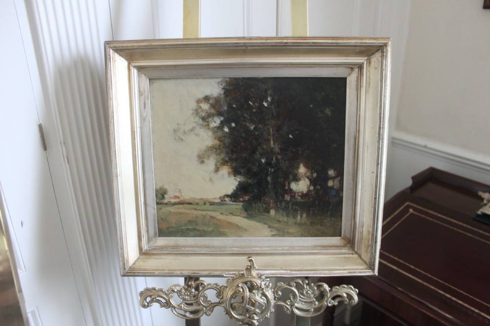 An antique oil painting on wooden panel depicting a muted rural landscape with trees and a track, presented in a wide silver gilt frame.