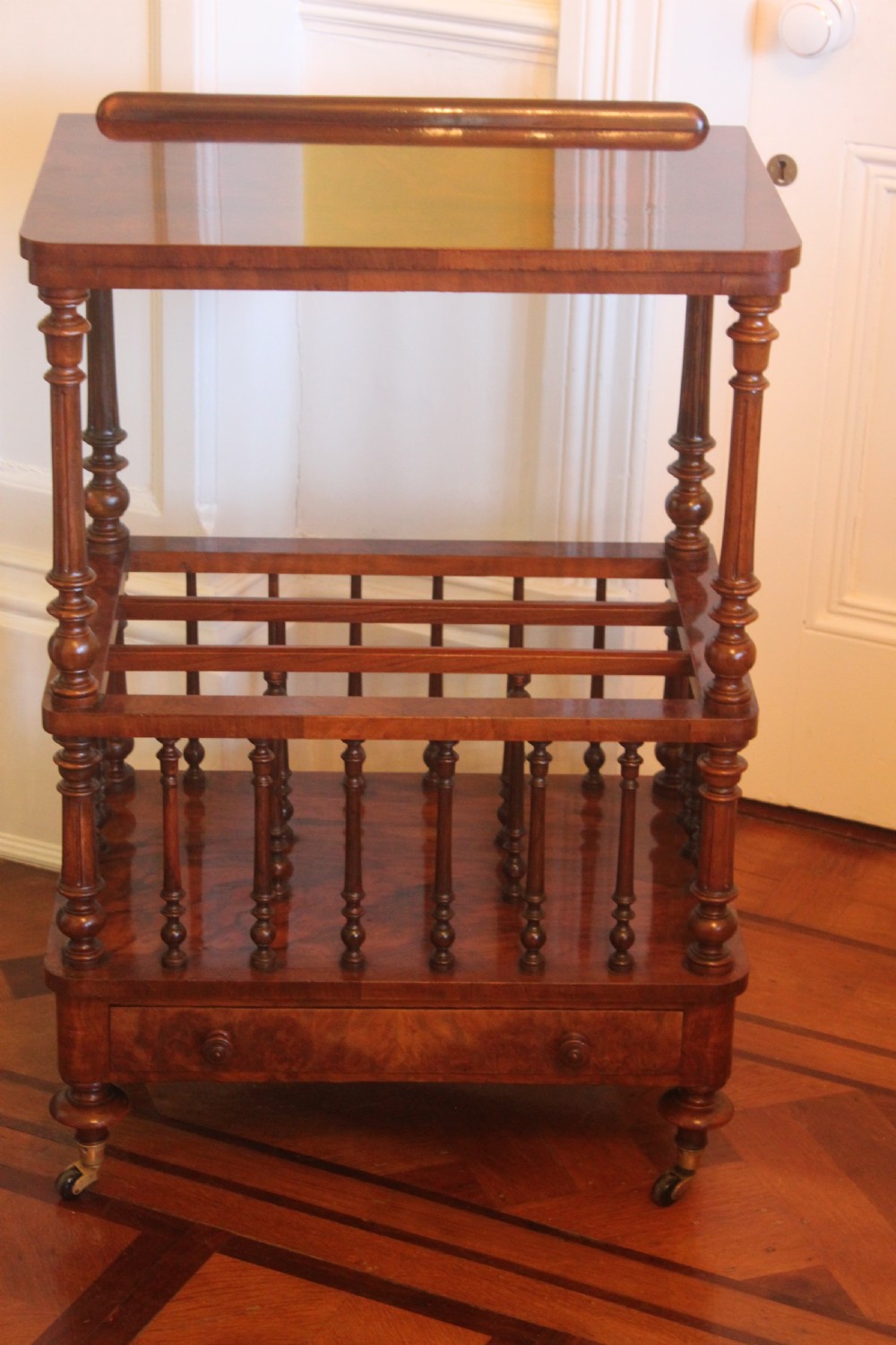 A Victorian burr walnut Canterbury what-not featuring a flat top surface with a gallery rail, turned spindle supports, a slatted middle partition section, a single base drawer, and turned legs with brass castors.