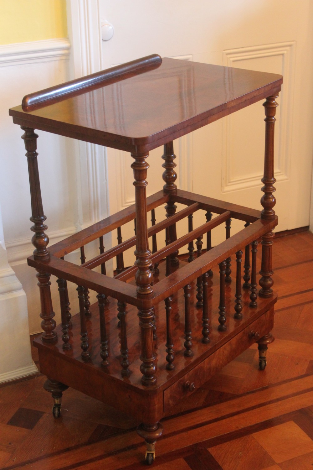 A Victorian burr walnut Canterbury what-not featuring a flat top surface with a gallery rail, turned spindle supports, a slatted middle partition section, a single base drawer, and turned legs with brass castors. View II