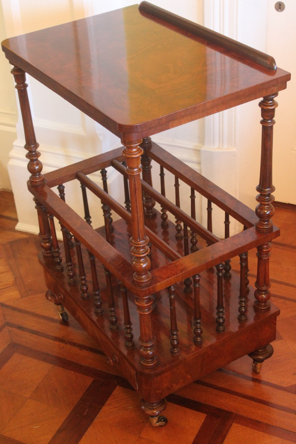 A Victorian burr walnut Canterbury what-not featuring a flat top surface with a gallery rail, turned spindle supports, a slatted middle partition section, a single base drawer, and turned legs with brass castors. View V