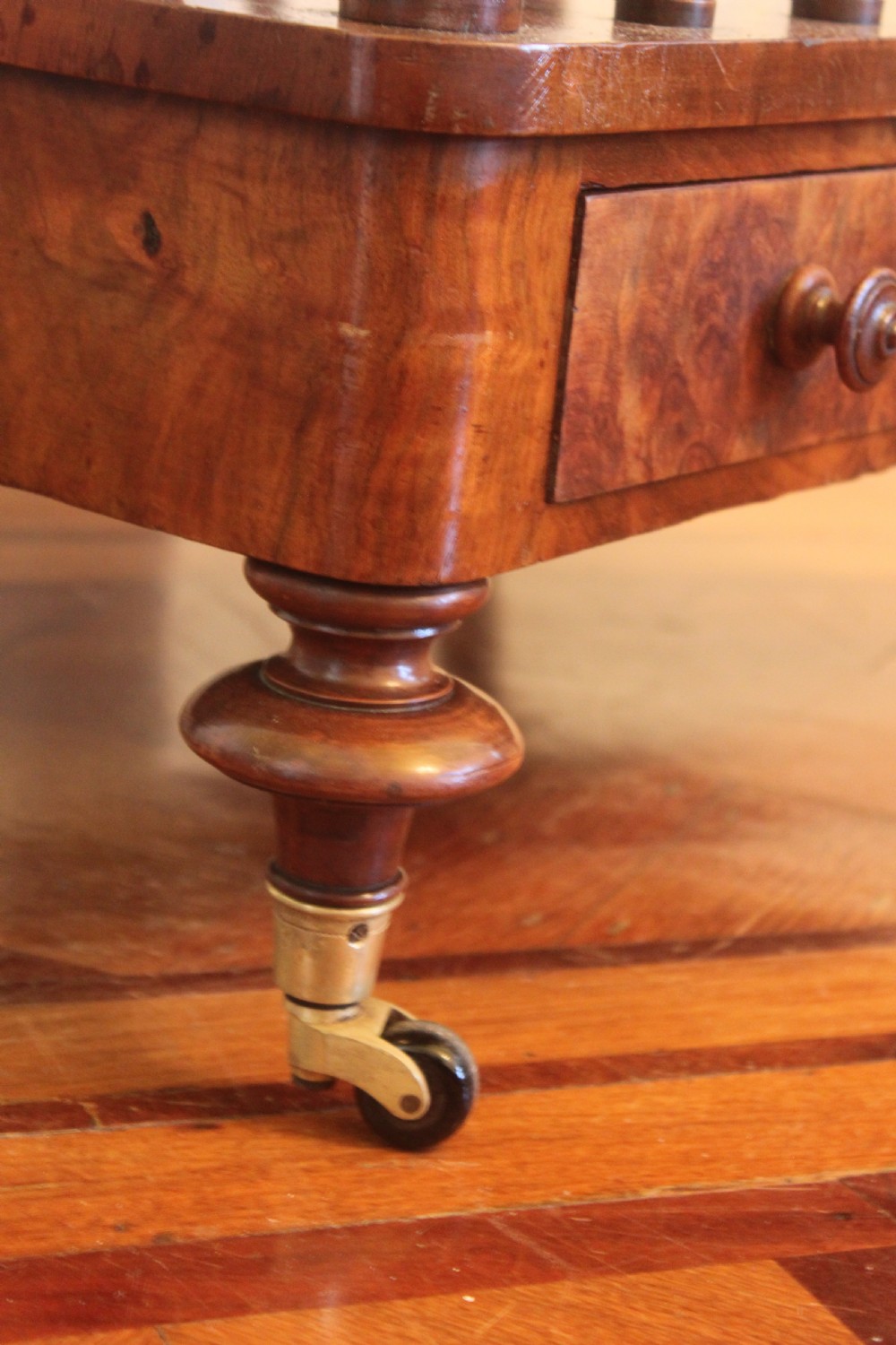 A Victorian burr walnut Canterbury what-not featuring a flat top surface with a gallery rail, turned spindle supports, a slatted middle partition section, a single base drawer, and turned legs with brass castors. View X
