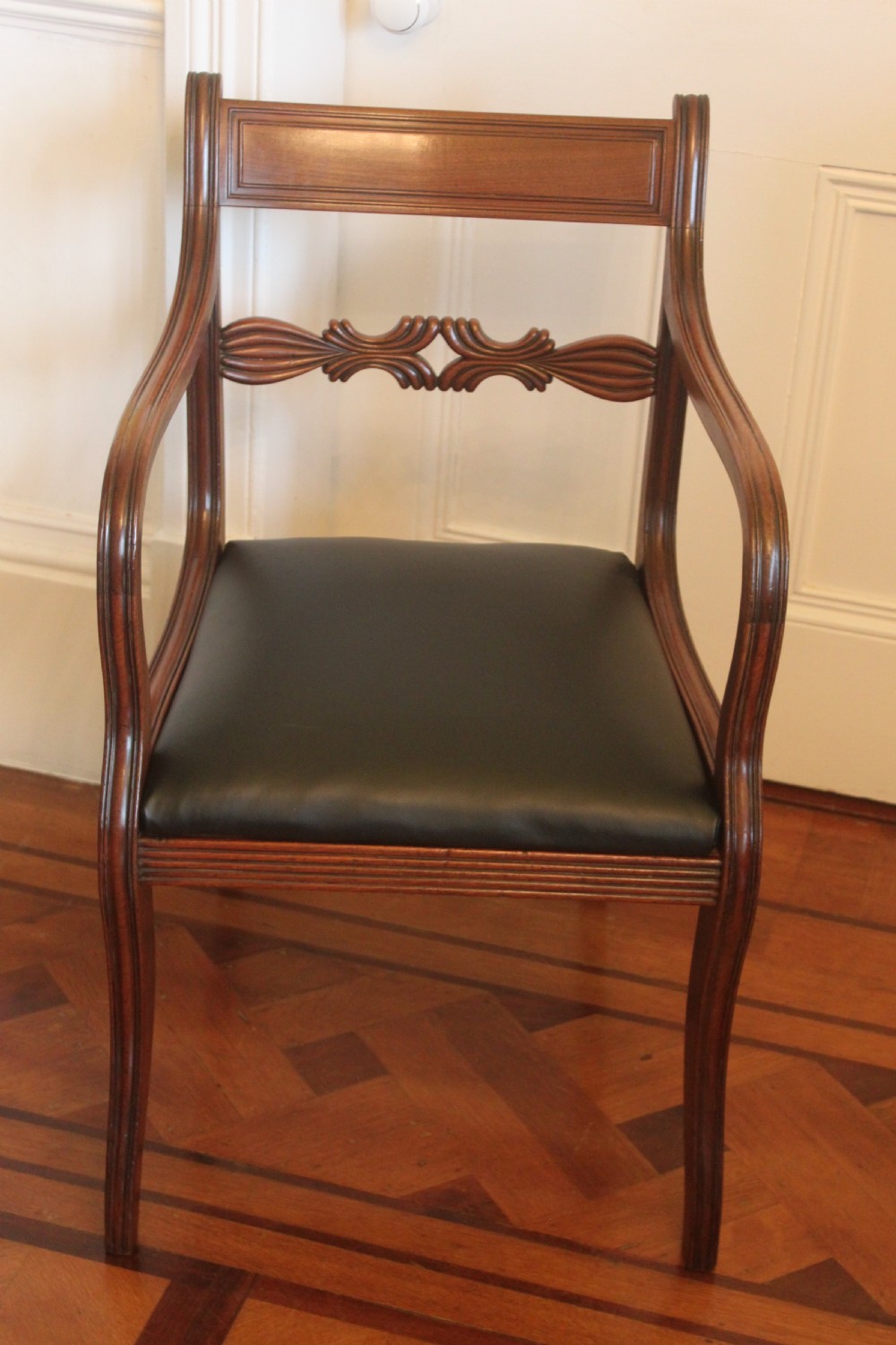 An antique Regency mahogany desk chair featuring sweeping reeded arms, a carved central back rail, and a black leather seat.