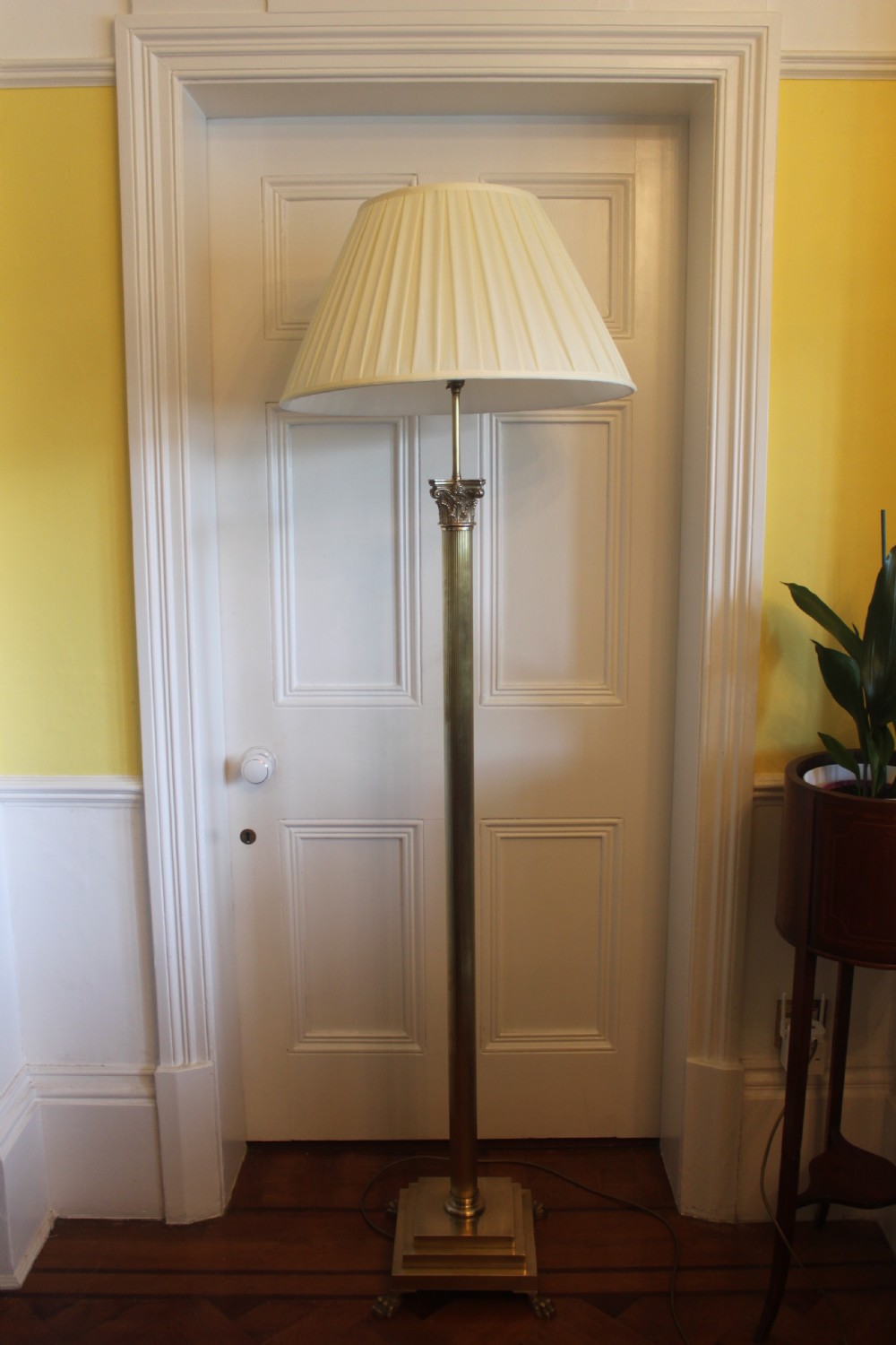 An antique brass standard lamp featuring a fluted column stem, decorative capital, stepped square pedestal base, and a cream pleated shade.