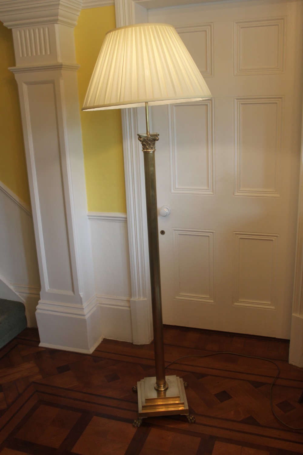 An antique brass standard lamp featuring a fluted column stem, decorative capital, stepped square pedestal base, and a cream pleated shade. View II