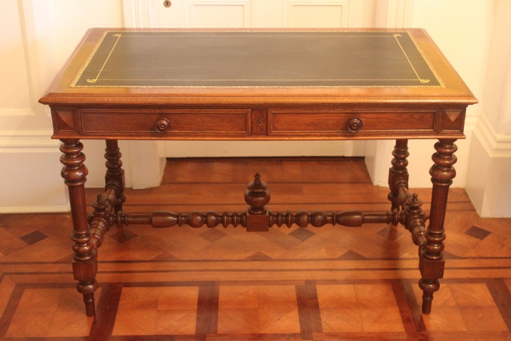 An antique French oak writing desk featuring a gold-tooled dark leather top insert, frieze drawers, turned baluster legs, and a stretcher base with a central finial.
