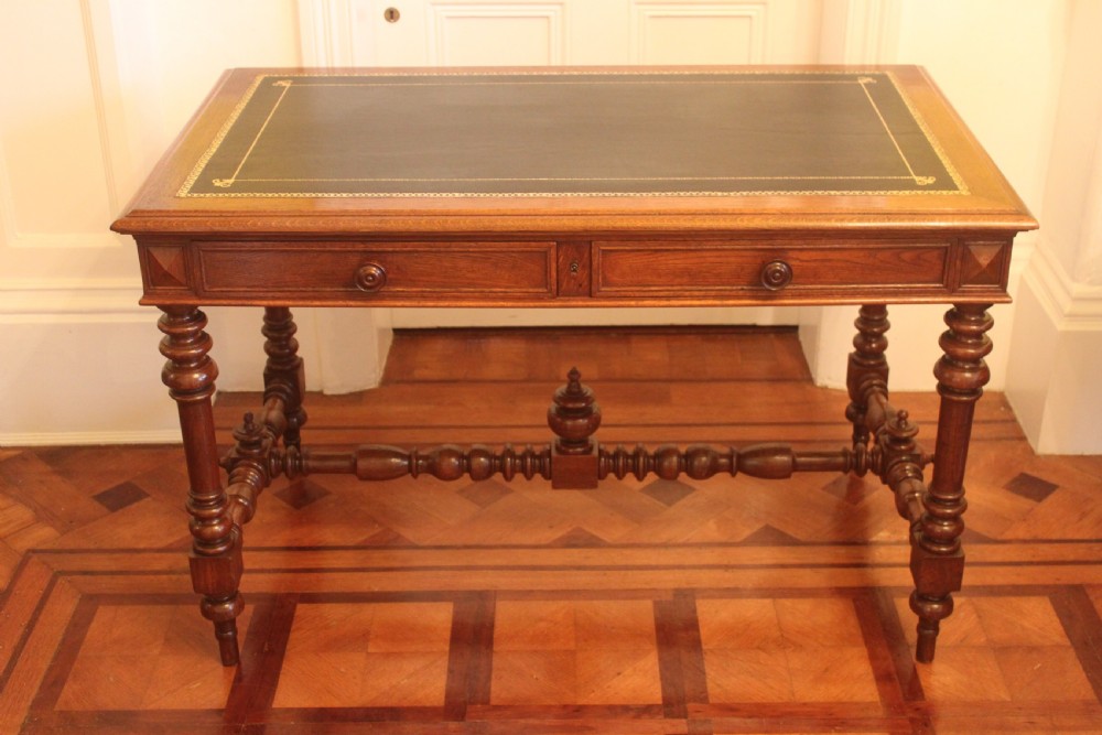 An antique French oak writing desk featuring a gold-tooled dark leather top insert, frieze drawers, turned baluster legs, and a stretcher base with a central finial. View II