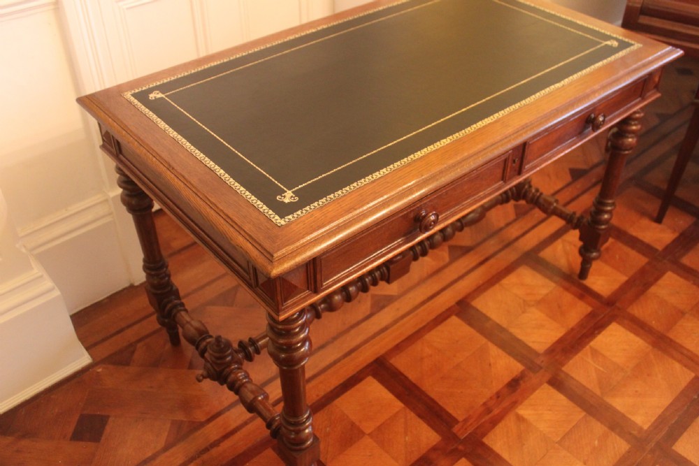 An antique French oak writing desk featuring a gold-tooled dark leather top insert, frieze drawers, turned baluster legs, and a stretcher base with a central finial. View V