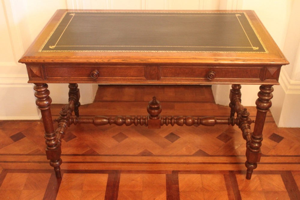 An antique French oak writing desk featuring a gold-tooled dark leather top insert, frieze drawers, turned baluster legs, and a stretcher base with a central finial. View VI