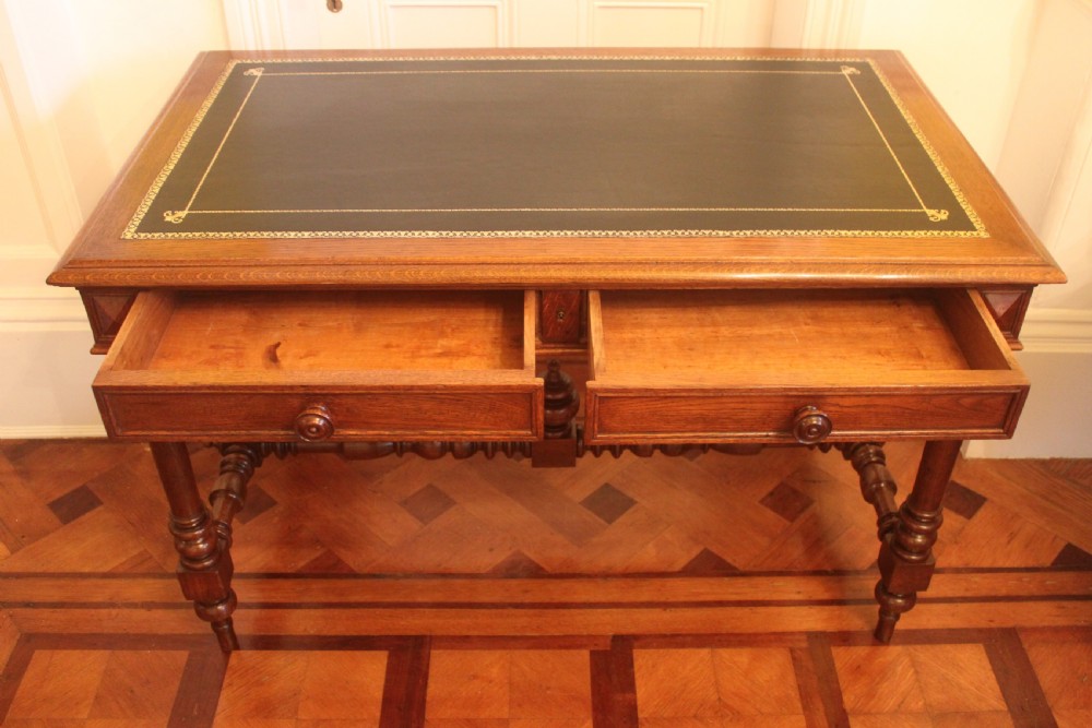 An antique French oak writing desk featuring a gold-tooled dark leather top insert, frieze drawers, turned baluster legs, and a stretcher base with a central finial. View X