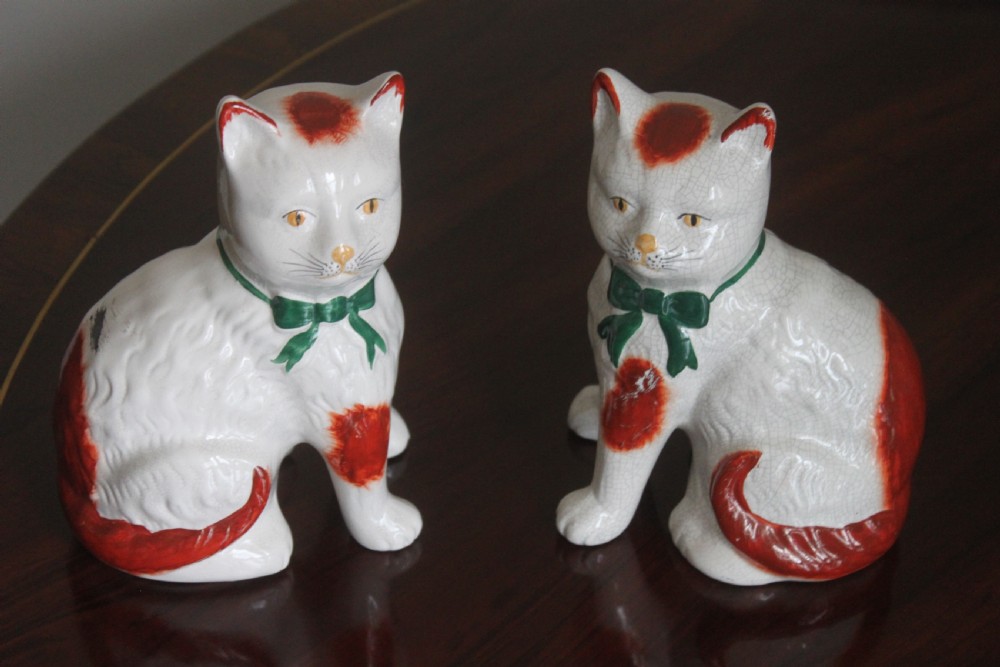 A pair of white press-moulded earthenware cat figurines featuring hand-painted rust-red patches and green ribbon bow collars.