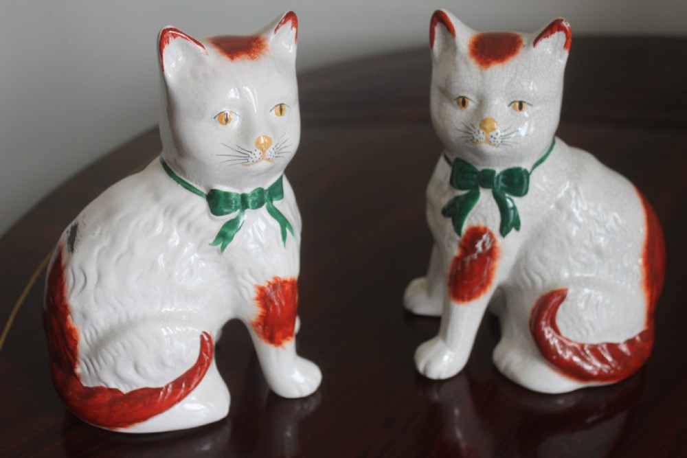 A pair of white press-moulded earthenware cat figurines featuring hand-painted rust-red patches and green ribbon bow collars. View II