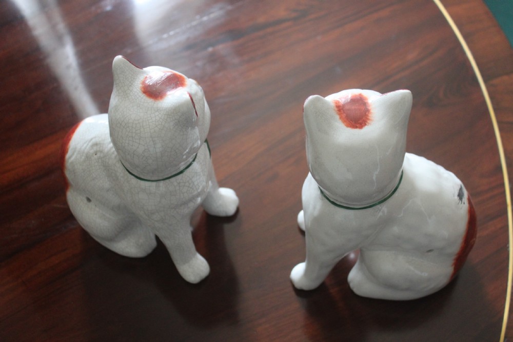 A pair of white press-moulded earthenware cat figurines featuring hand-painted rust-red patches and green ribbon bow collars. View V