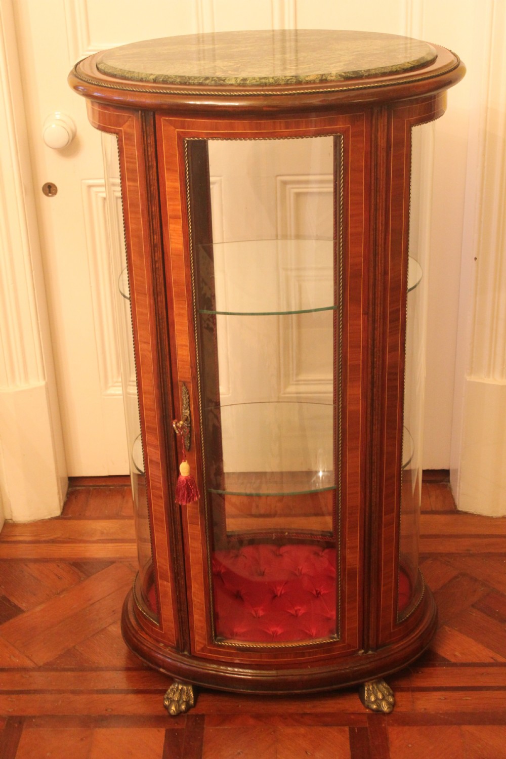 An oval antique French vitrine cabinet featuring an inset green marble top, inlaid timber marquetry, curved glass panels, and brass claw feet.