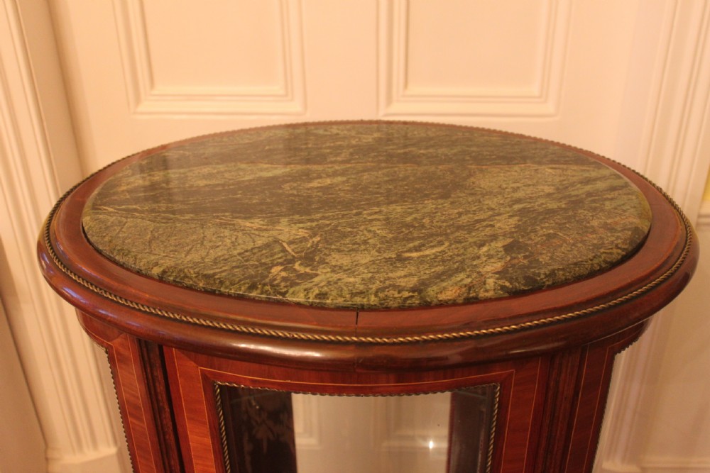 An oval antique French vitrine cabinet featuring an inset green marble top, inlaid timber marquetry, curved glass panels, and brass claw feet. View V
