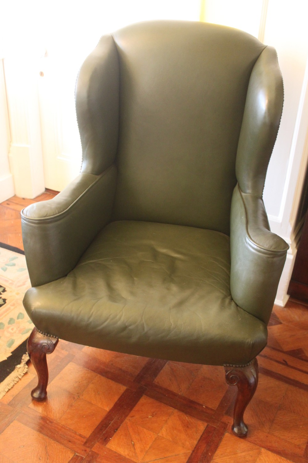 A late nineteenth or early twentieth century Queen Anne style wingback armchair upholstered in olive green leather with carved cabriole legs.