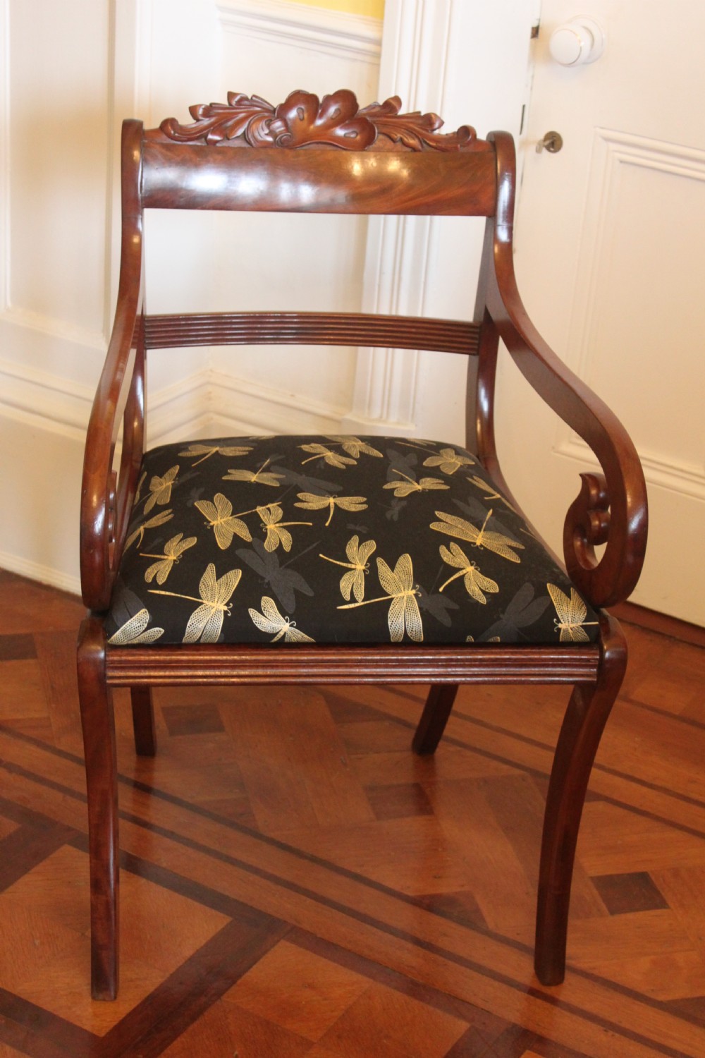 A Regency mahogany armchair featuring a carved crest rail, scrolled reeded arms, sabre front legs, and dragonfly pattern upholstery.