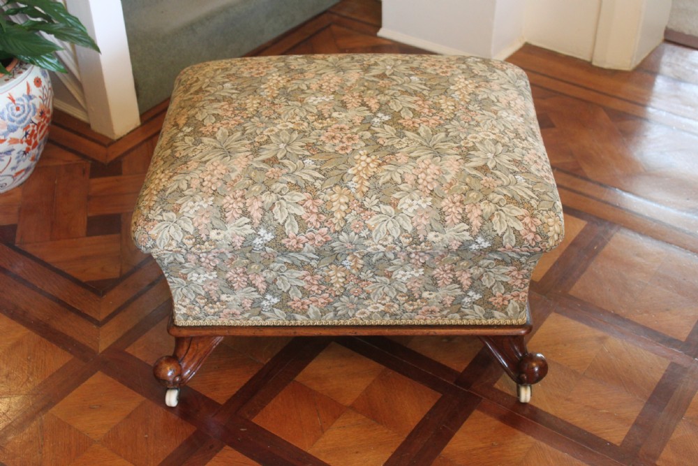 A square Victorian mahogany footstool upholstered in a beige, pink, and green floral tapestry pattern, supported by carved scroll legs with white ceramic castors.