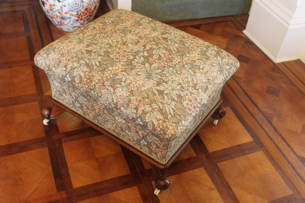 A square Victorian mahogany footstool upholstered in a beige, pink, and green floral tapestry pattern, supported by carved scroll legs with white ceramic castors. View II