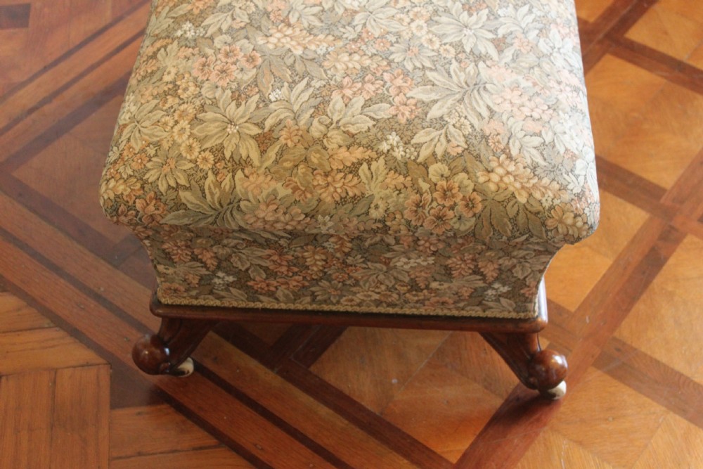 A square Victorian mahogany footstool upholstered in a beige, pink, and green floral tapestry pattern, supported by carved scroll legs with white ceramic castors. View III