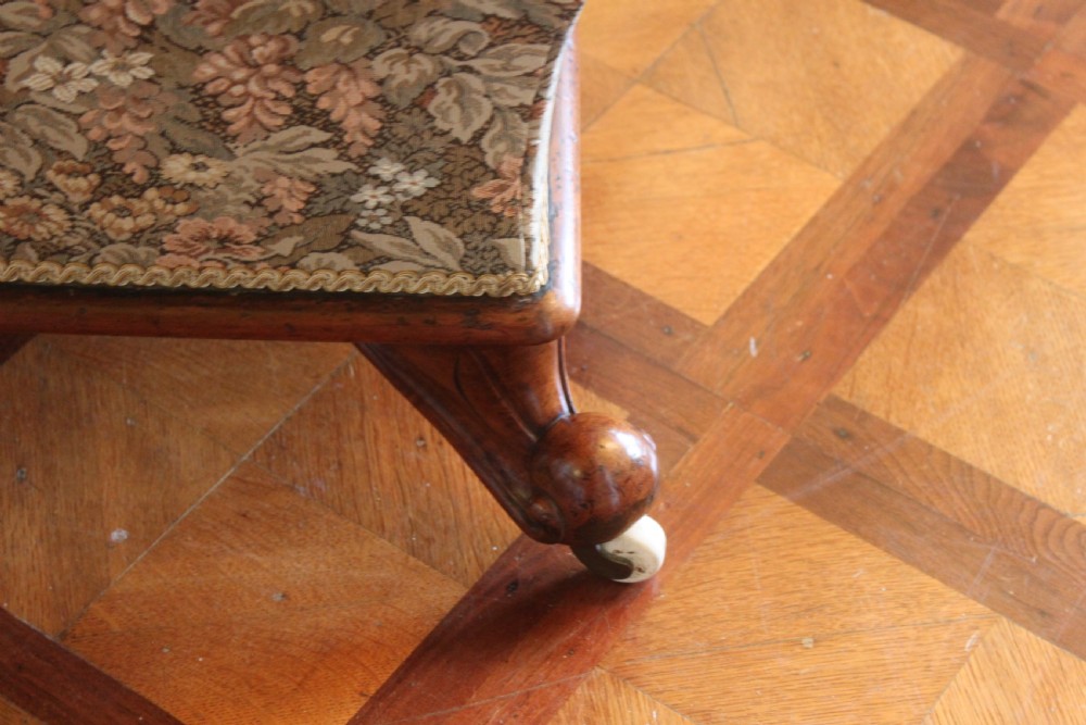 A square Victorian mahogany footstool upholstered in a beige, pink, and green floral tapestry pattern, supported by carved scroll legs with white ceramic castors. View V