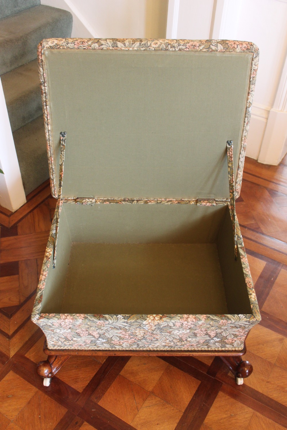 A square Victorian mahogany footstool upholstered in a beige, pink, and green floral tapestry pattern, supported by carved scroll legs with white ceramic castors. View VI