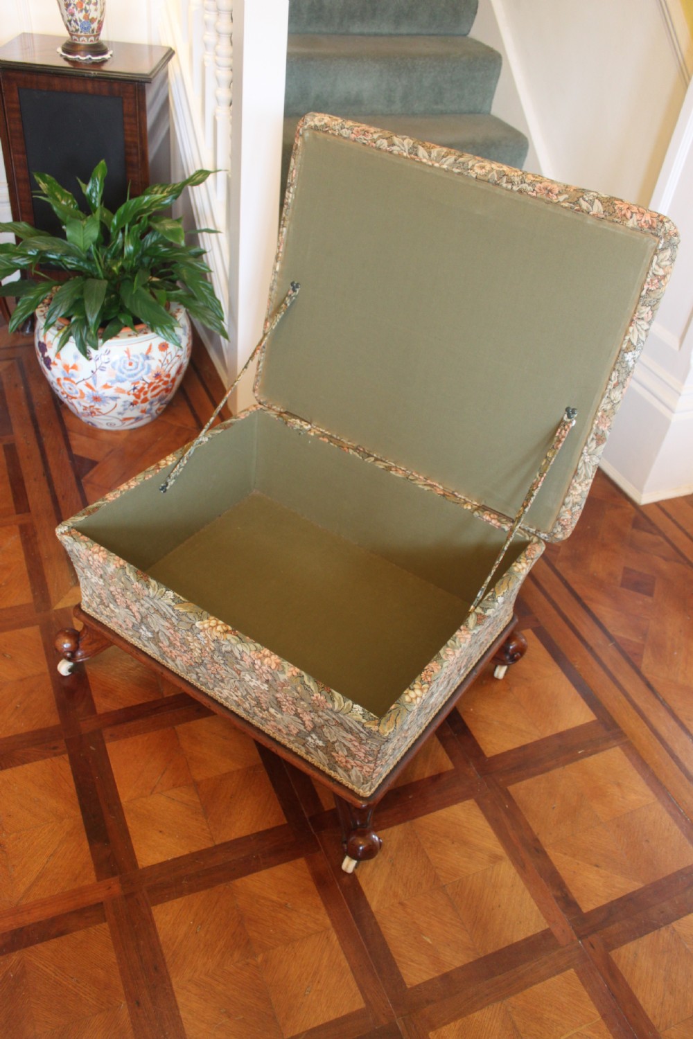 A square Victorian mahogany footstool upholstered in a beige, pink, and green floral tapestry pattern, supported by carved scroll legs with white ceramic castors. View VII