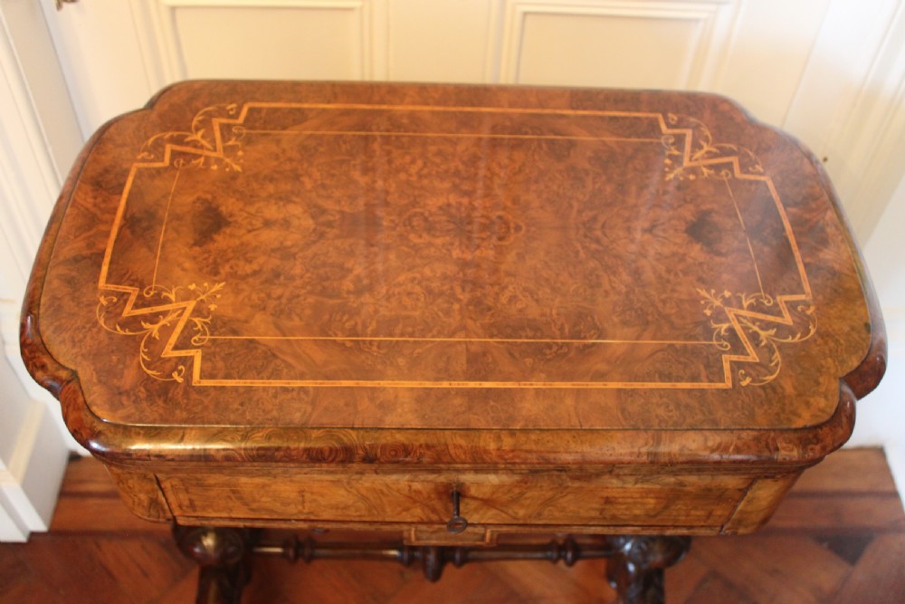 A Victorian burr walnut games and sewing table featuring inlay ornamentation, turned column supports, a wooden work well, and carved legs on castors. View II