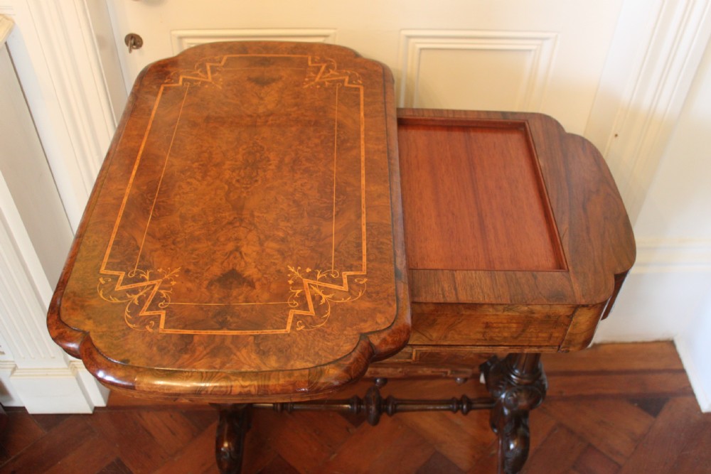 A Victorian burr walnut games and sewing table featuring inlay ornamentation, turned column supports, a wooden work well, and carved legs on castors. View V