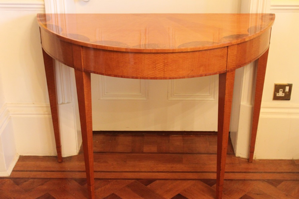 An early 19th-century semi-circular satinwood demi-lune console table featuring decorative top marquetry and four square tapered legs.