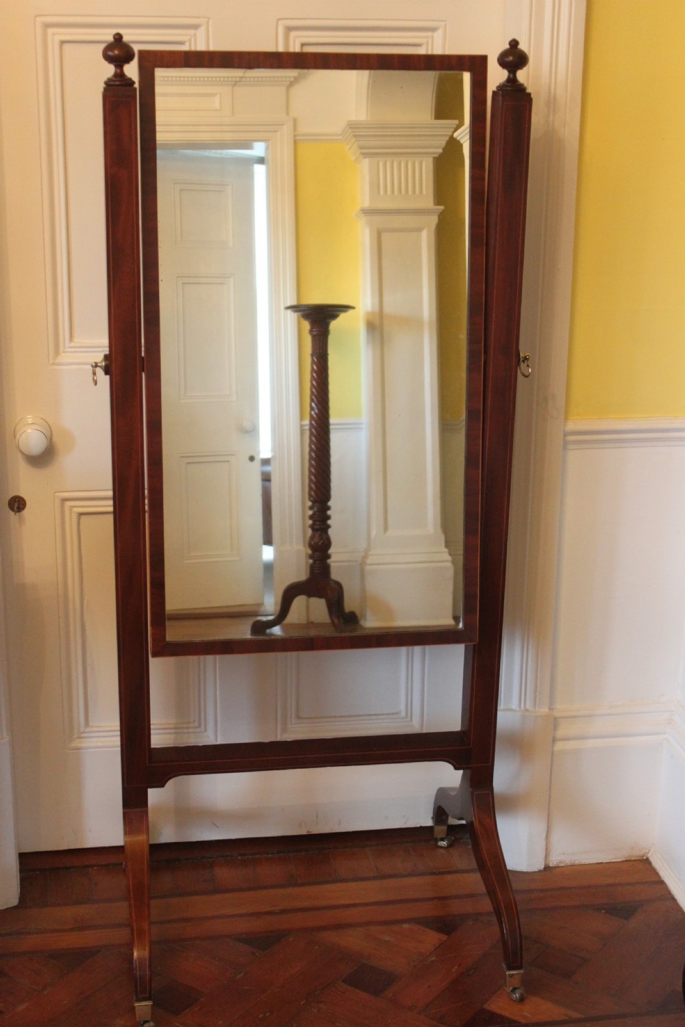 A full-length Sheraton style mahogany cheval dressing mirror with turned finials, splayed legs, brass castors, and a tilting frame.