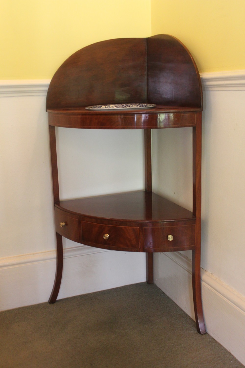 A Georgian mahogany corner washstand featuring an upper arched splashback, a central tier with a drawer, brass knobs, and curved splayed legs.
