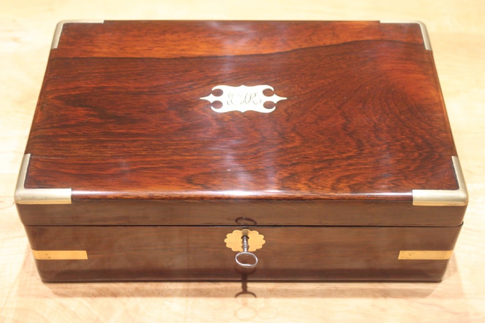 A rectangular Victorian rosewood writing box featuring flush brass corner brackets, a central engraved brass cartouche, and a key inserted into the front keyhole.