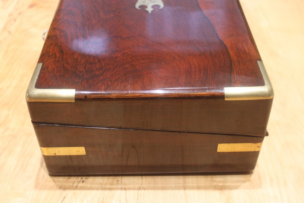 A rectangular Victorian rosewood writing box featuring flush brass corner brackets, a central engraved brass cartouche, and a key inserted into the front keyhole. View V