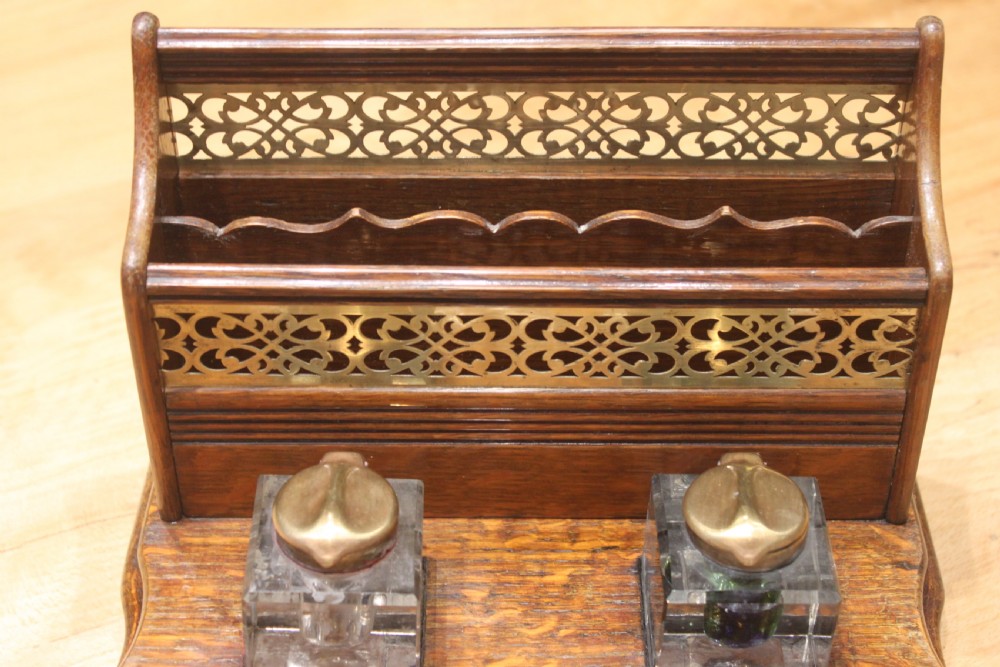 Late Victorian oak standish featuring a stationary gallery with pierced brass trim, two square glass inkwells with brass lids, and a carved pen groove. View II
