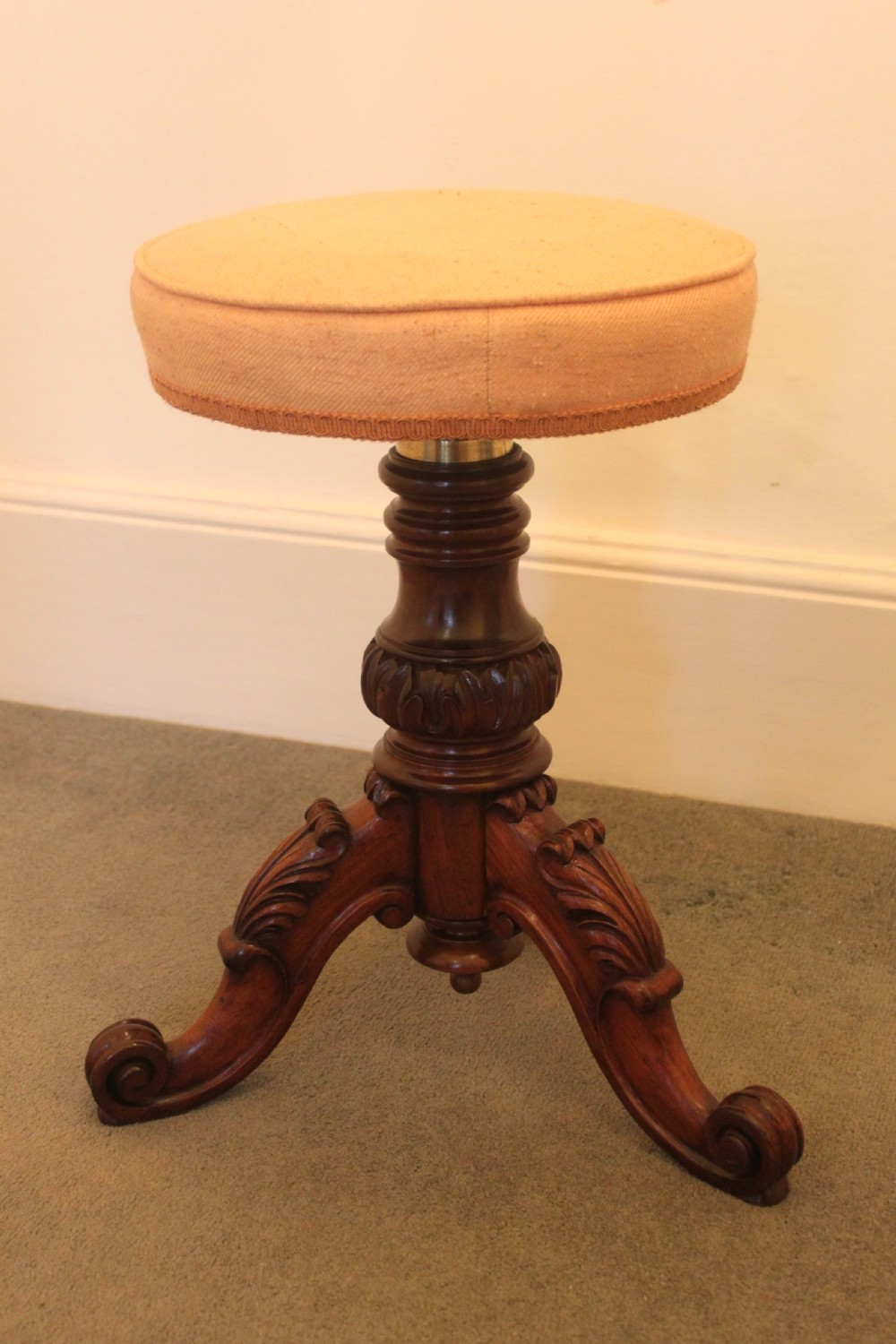 A Victorian mahogany revolving piano stool featuring a circular beige upholstered seat, a turned pedestal with acanthus leaf carving, and tripod scroll feet.