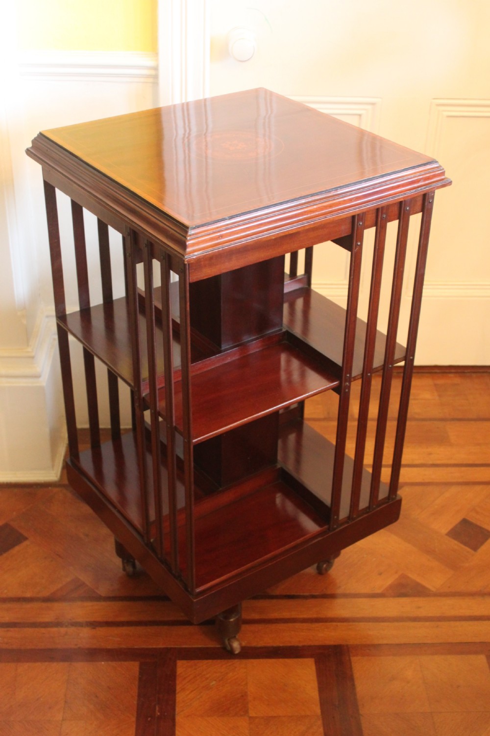 An Edwardian mahogany revolving bookcase featuring a central satinwood marquetry inlay top, slatted sides, open shelves, and castors. View II