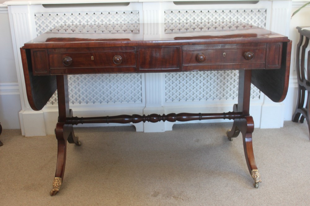 An early nineteenth-century mahogany sofa table featuring two drop leaves, two frieze drawers with knob handles, a turned central stretcher, and splayed legs with brass paw feet and castors.
