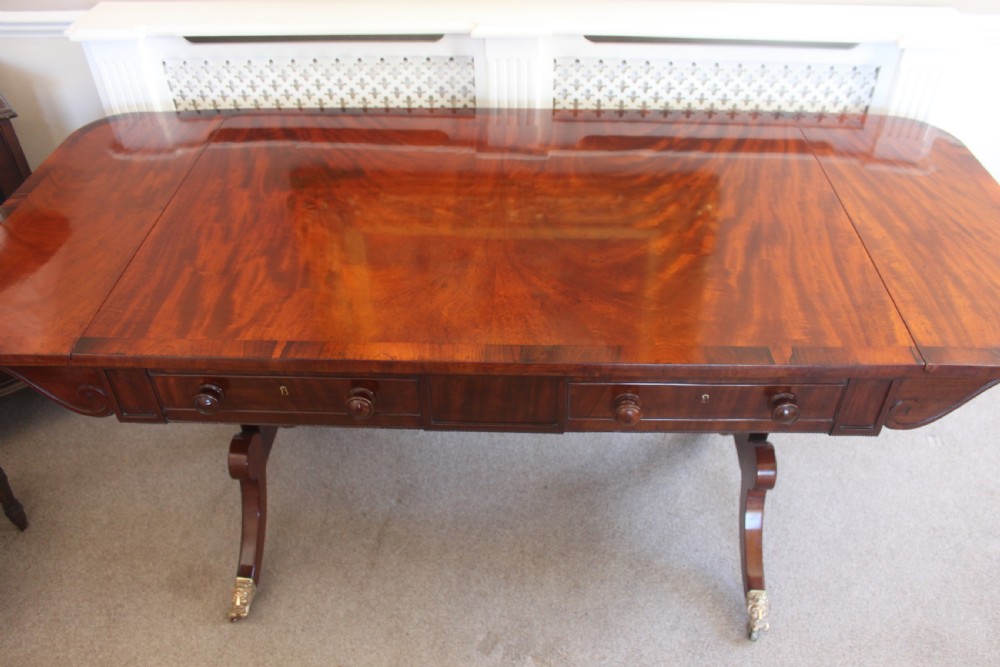 An early nineteenth-century mahogany sofa table featuring two drop leaves, two frieze drawers with knob handles, a turned central stretcher, and splayed legs with brass paw feet and castors. View VII