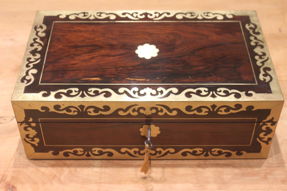 A rectangular dark rosewood box decorated with intricate brass scrollwork inlay along the borders and top centre, complete with a key inserted into the front lock.