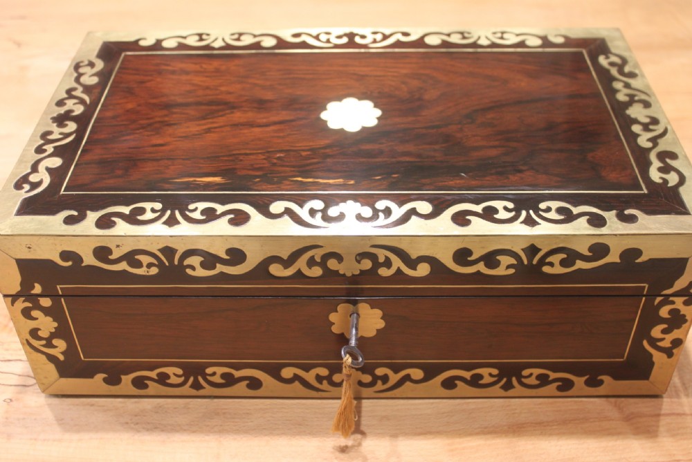 A rectangular dark rosewood box decorated with intricate brass scrollwork inlay along the borders and top centre, complete with a key inserted into the front lock. View II