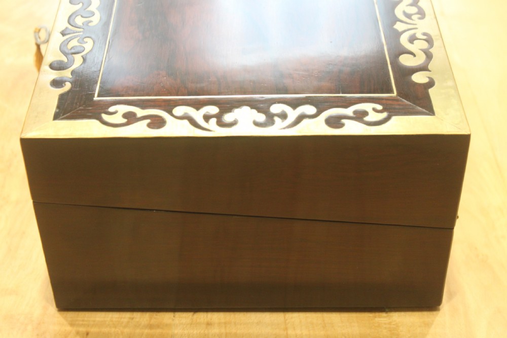 A rectangular dark rosewood box decorated with intricate brass scrollwork inlay along the borders and top centre, complete with a key inserted into the front lock. View V