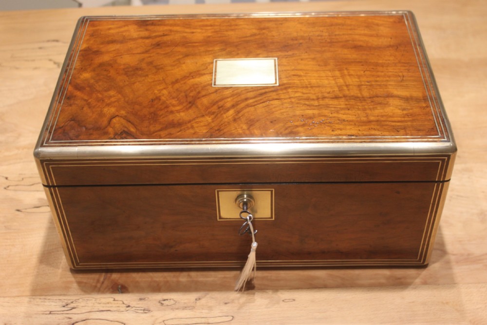 A closed Victorian writing slope crafted from figured walnut veneer featuring brass edging, line inlay, a brass cartouche, and a key inserted in the Bramah lock with a tassel.