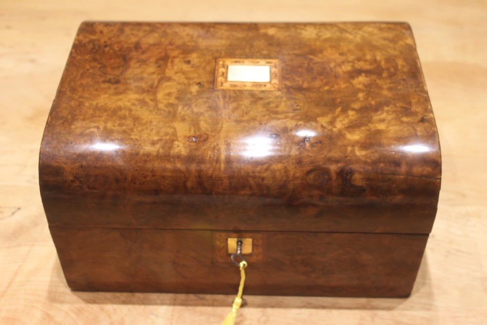 A rectangular Victorian burr walnut writing slope box featuring a domed lid, central inlaid cartouche, and a key inserted into the brass escutcheon.