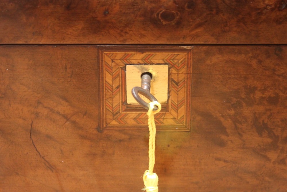 A rectangular Victorian burr walnut writing slope box featuring a domed lid, central inlaid cartouche, and a key inserted into the brass escutcheon. View V